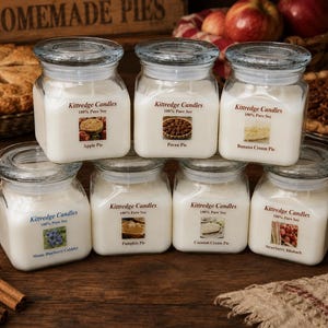 May include: Seven square glass jars filled with white soy wax candles, each featuring a Kittredge Candles label with a specific pie or dessert image and name, including Apple Pie, Pecan Pie, Banana Cream Pie, Maine Blueberry Cobbler, Pumpkin Pie, Coconut Cream Pie, and Strawberry Rhubarb.