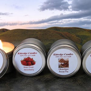 May include: Four round silver metal tins containing soy candles. Each lid features a Kittredge Candles label with a unique image and text: Waving Wheat, State Fruit: Strawberry, Okmulgee Pecan Pie, and Oklahoma Rose. One candle is lit.
