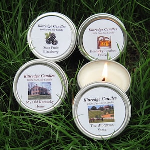 May include: Five small, round, silver-colored candle tins with white labels and various images and text, including "State Fruit: Blackberry" and "Kentucky Bourbon Festival." One candle is lit, with a visible flame. The candles are set on green grass.