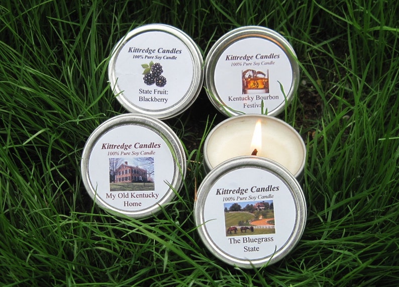 May include: Five small, round, silver-colored candle tins with white labels and various images and text, including "State Fruit: Blackberry" and "Kentucky Bourbon Festival." One candle is lit, with a visible flame. The candles are set on green grass.