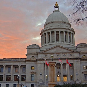 May include: The Arkansas State Capitol building features a large central dome with a gold-colored finial, neoclassical stone architecture, and multiple columns. Three flags, including the American flag, fly in front under a sunset sky.