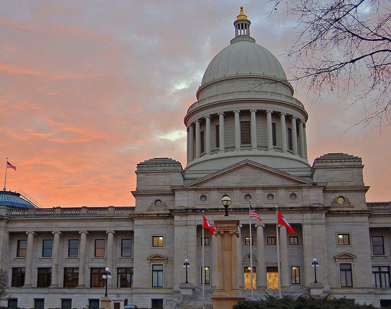 May include: The Arkansas State Capitol building features a large central dome with a gold-colored finial, neoclassical stone architecture, and multiple columns. Three flags, including the American flag, fly in front under a sunset sky.