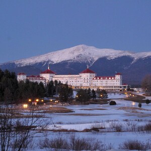 May include: A large white hotel building with a red roof sits in front of a snow-covered mountain range during twilight. The structure features multiple stories, numerous windows, and illuminated lights, surrounded by a snowy landscape and trees.