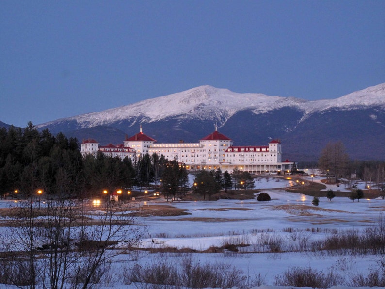 May include: A large white hotel building with a red roof sits in front of a snow-covered mountain range during twilight. The structure features multiple stories, numerous windows, and illuminated lights, surrounded by a snowy landscape and trees.
