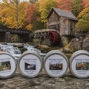 May include: Four round silver metal candle tins with white labels featuring the text Kittredge Candles 100% Pure Soy Candle and specific scent names. One candle is lit with a visible flame, while the others are closed with their lids.