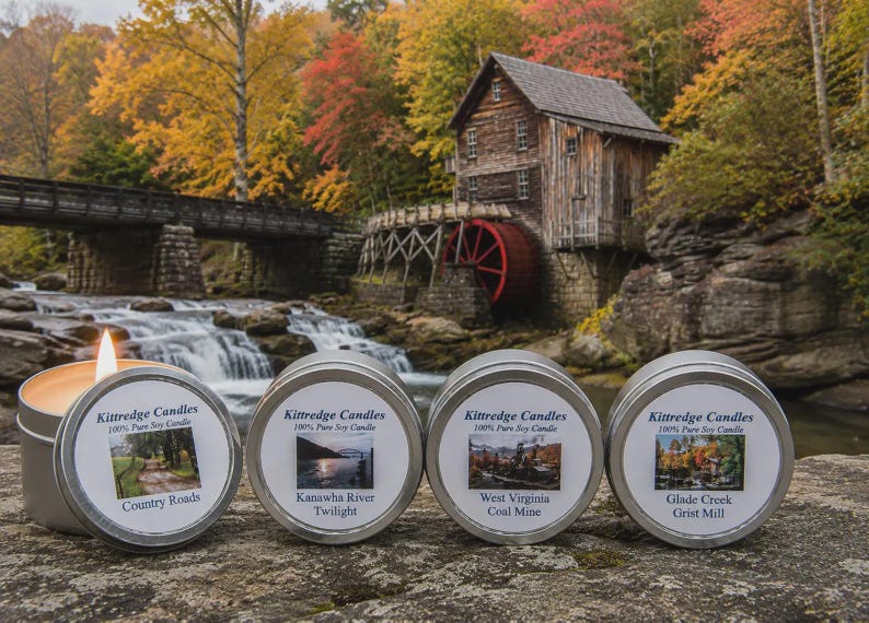May include: Four round silver metal candle tins with white labels featuring the text Kittredge Candles 100% Pure Soy Candle and specific scent names. One candle is lit with a visible flame, while the others are closed with their lids.
