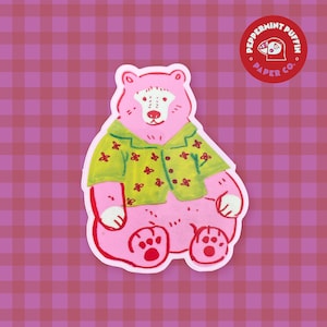 May include: A pink bear sticker wearing a green shirt with red flower accents. The sticker has a white border and is on a pink and red plaid background. A circular logo with the text "Peppermint Puffin Paper Co." is in the upper right corner.