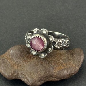 May include: A silver ring featuring a central faceted round pink gemstone set in a serrated bezel. The ring band is shaped like a flower with petals surrounding the stone and has a textured, floral-patterned shank with an oxidized finish.