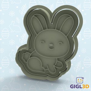 Puede incluir: Un cortador de galletas de plástico gris con forma de conejito. El cortador presenta un conejito sonriente con orejas largas y una cola esponjosa. El cortador está diseñado con múltiples capas para formas de galletas detalladas. El fondo es azul claro con patrones de Pascua.