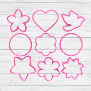 Puede incluir: Nueve cortadores de galletas rosas en varias formas, incluyendo flores, un corazón, una paloma, un círculo, una nube, un círculo festoneado, una estrella y dos diseños florales más. Los cortadores están dispuestos sobre una superficie de madera blanca.