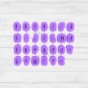 May include: A set of purple alphabet letter cookie cutters arranged on a white wooden surface. The letters are in a rounded, sans-serif font. Each letter is connected to a handle for easy use.