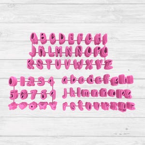 Pode incluir: Cortadores de biscoitos rosa de plástico com letras e números. O conjunto inclui letras maiúsculas e minúsculas, números de 0 a 9 e várias formas decorativas pequenas. Os cortadores estão dispostos sobre uma superfície de madeira branca.