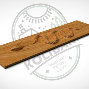 Könnte beinhalten: Rechteckige Holzplakette mit einem geschnitzten Schlangenmuster. Die Schlange ist in einer gewundenen Position dargestellt, mit detaillierten Schuppen und einer strukturierten Oberfläche. Das Holz hat eine natürliche, hellbraune Farbe. Der Text "KOLIBAR" und "Wood work that Tells a Story" sind sichtbar.