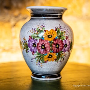 Pode incluir: Um vaso de cerâmica branco com um desenho floral. O vaso tem um corpo redondo e um pescoço estreito. As flores são pintadas em tons de roxo, amarelo e vermelho.