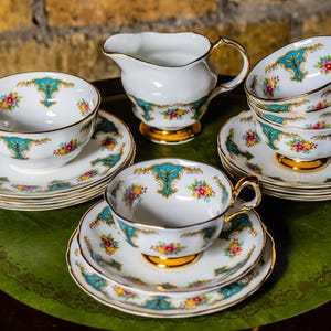 Può includere: Un set di tazze da tè e piattini vintage con un design floreale e turchese. Le tazze e i piattini sono bianchi con bordi dorati.