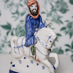 Op de afbeelding: Een keramieken beeldje dat een man op een wit paard voorstelt. De man draagt een blauwe jurk en een rode tulband. Het paard is versierd met blauwe accenten. De basis is blauw met bloemmotieven.