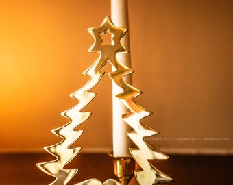 May include: A gold-coloured Christmas tree candle holder with a lit white candle. The tree-shaped holder has a star at the top and zig-zag edges. The candle holder is on a wooden surface.