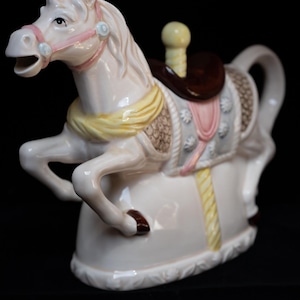 Op de afbeelding: Een witte keramische theepot in de vorm van een draaimolenpaard met een bruin zadel, gele en roze details en een gele touwgreep.