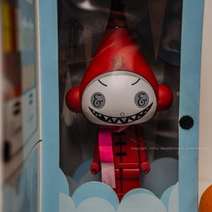 Könnte beinhalten: Rote Figur mit spitzem Hut und weißem Gesicht mit breitem, zahnigem Grinsen. Die Figur hat einen rosa Schal und wird in einer hellblauen Box mit Wolkenmuster am Boden präsentiert. Die Zahl "13" ist auf dem Hut sichtbar.