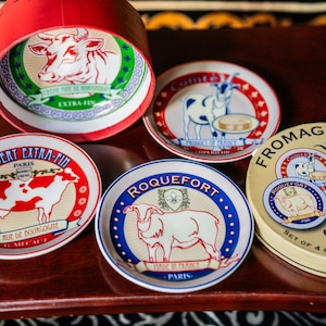 May include: A set of four small round ceramic cheese plates featuring illustrations of cows, goats, and sheep, with labels including Camembert Extra-Fin, Roquefort, and Comté. The plates are presented in a round cream-coloured box with red accents.