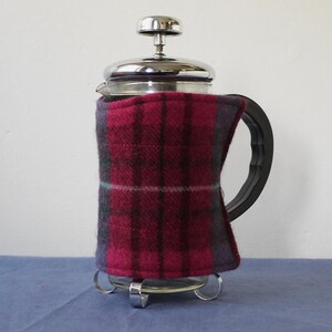 May include: A French press coffee maker with a stainless steel lid and base. The glass carafe is wrapped in a red, grey, and teal plaid cosy. The black handle is on the right side of the carafe.