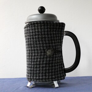 May include: A black and white checked fabric coffee pot cosy with a black button. The cosy is on a glass coffee pot with a black lid.