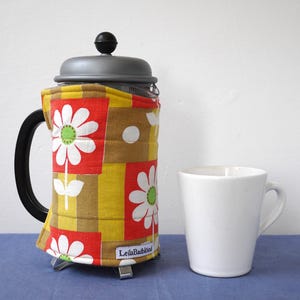 May include: A French press coffee maker with a floral patterned cosy. The cosy has a patchwork design with white daisies on red, yellow, and brown squares. A white mug is next to the coffee maker. The text "LeilaBadblood" is on the cosy.