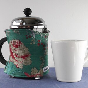 May include: A French press coffee maker with a teal floral cosy and a white ceramic mug. The cosy features a large red and cream floral pattern. The coffee maker has a black handle and a silver lid.