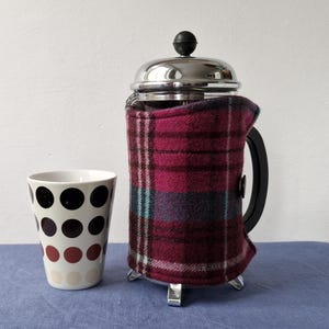 May include: A French press coffee maker with a burgundy plaid cosy, and a white ceramic mug with polka dots. The coffee maker has a black handle and stainless steel accents. The items are on a blue surface.