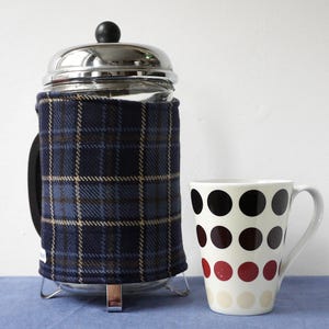 May include: A French press coffee maker with a navy blue plaid cosy and a white mug with black, burgundy, and cream polka dots. The coffee maker has a stainless steel lid and base.
