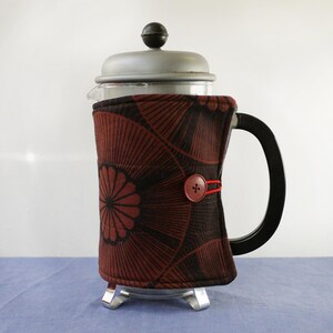 May include: A French press coffee maker with a black handle and a patterned fabric cosy. The cosy is dark red and black with a button closure. The lid and base are silver.