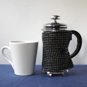 May include: A black and white patterned fabric coffee press with a glass carafe and a chrome lid. The coffee press is sitting on a silver stand.