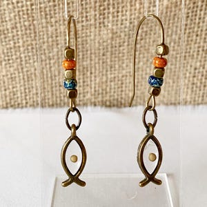 May include: A pair of handcrafted earrings featuring a fish symbol. The earrings have a gold-colored hook and are adorned with colorful beads in orange, blue, and gold. The fish pendant has a white center with a small, light-colored dot.