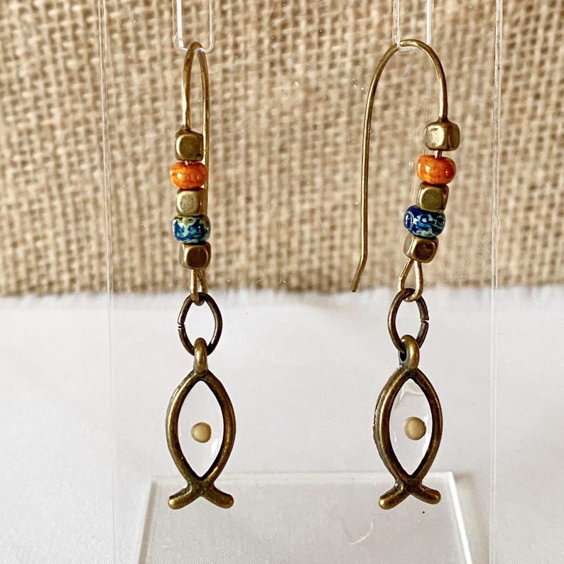 May include: A pair of handcrafted earrings featuring a fish symbol. The earrings have a gold-colored hook and are adorned with colorful beads in orange, blue, and gold. The fish pendant has a white center with a small, light-colored dot.