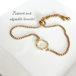 May include: A gold-coloured adjustable bracelet featuring a mustard seed in a clear oval pendant. The delicate chain is visible, and the text "Mustard seed adjustable bracelet" is present.
