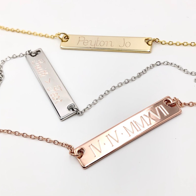 May include: Three gold bar necklaces with different engravings. The top necklace has the name "Peyton Jo" engraved on the bar. The middle necklace has the words "Smith & Beck" engraved on the bar. The bottom necklace has the Roman numerals "IV.IV.MMXVI" engraved on the bar.