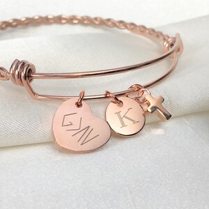 May include: Rose gold bangle bracelet with three charms: a heart with the inscription "G.W.N." a round charm with the letter "K" and a small cross charm.