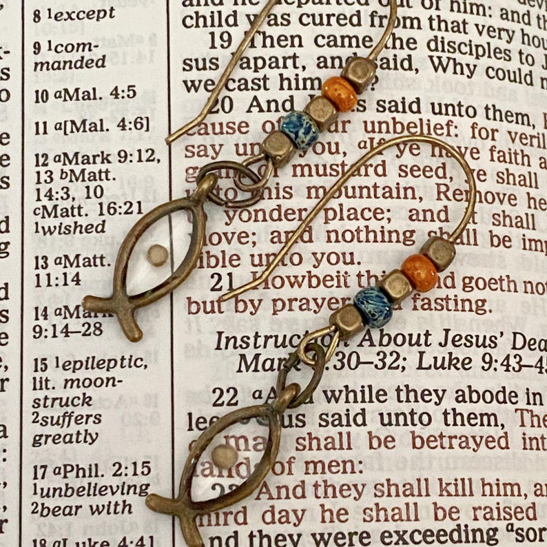 May include: A pair of handcrafted earrings featuring a fish symbol with a small bead inside. The earrings have a bronze finish and are accented with colorful beads in shades of blue, orange, and gold. The earrings are displayed against a backdrop of aged paper.