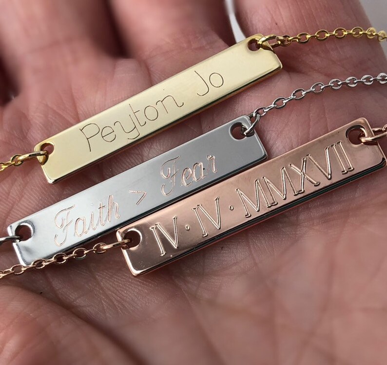 May include: Three gold bar necklaces with different engravings. The top necklace has the name "Peyton Jo" engraved on it. The middle necklace has the words "Faith > Fear" engraved on it. The bottom necklace has the Roman numerals "IV.IV.MMXVI" engraved on it.