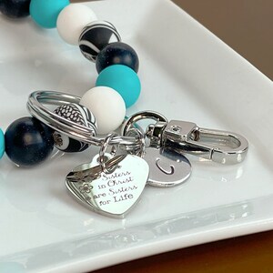 May include: A silver keychain with a heart charm that reads "Sisters in Christ are Sisters for Life". The keychain also has a silver letter "C" charm and a silver clasp. The keychain is attached to a beaded bracelet with black, white, and turquoise beads.