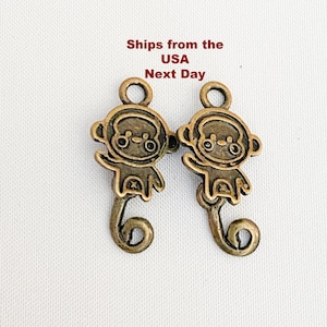 Op de afbeelding: Twee bronzen aapjesbedels met een lus om op te hangen. De bedels hebben een gedetailleerd ontwerp, inclusief een apengezicht, armen en een gekrulde staart. De tekst "Ships from the USA Next Day" is zichtbaar bovenaan.