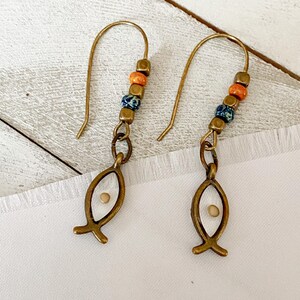 May include: A pair of brass-colored earrings. Each earring features a fish-shaped charm with a small seed inside, connected to a beaded section with orange, blue, and brown beads. The earrings have a hook closure.