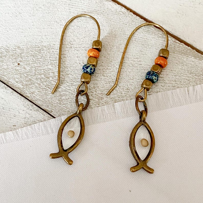May include: A pair of brass-colored earrings. Each earring features a fish-shaped charm with a small seed inside, connected to a beaded section with orange, blue, and brown beads. The earrings have a hook closure.