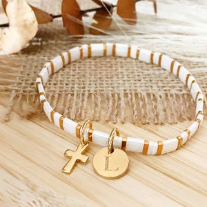 Puede incluir: Pulsera de cuentas blancas y doradas con un dije de cruz dorada y un dije circular dorado con la letra "L". La pulsera está sobre una superficie de madera con un fondo de arpillera.