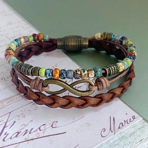 May include: A multi-strand leather bracelet with a colorful beaded row and an infinity symbol charm. The bracelet has a brown leather strap with a braided design.