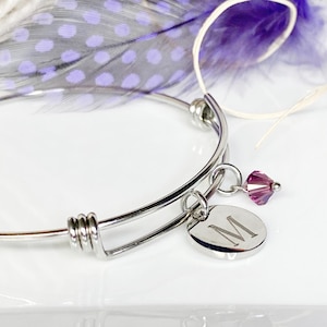 May include: Expandable silver bracelet with a round charm featuring the letter "M" and a small, faceted, purple crystal bead. The bracelet is displayed with a purple and white feather.