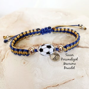 May include: A blue and gold macrame bracelet with a football charm and a silver letter A charm. The bracelet is tied with a gold cord and has silver beads. Soccer Personalised Macrame Bracelet.
