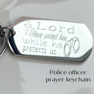 May include: Silver keychain with a cross and handcuffs engraved with the text "Lord, Please protect him while he protects us."