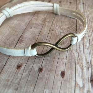 May include: A white leather bracelet with an infinity symbol charm. The charm is made of brass and has a polished finish.