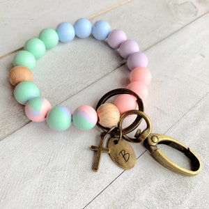 Op de afbeelding: Een pastelkleurige siliconen kralen armband met een houten kraal en een gouden sleutelhanger met een kruisbedel en een letter 'B' bedel.